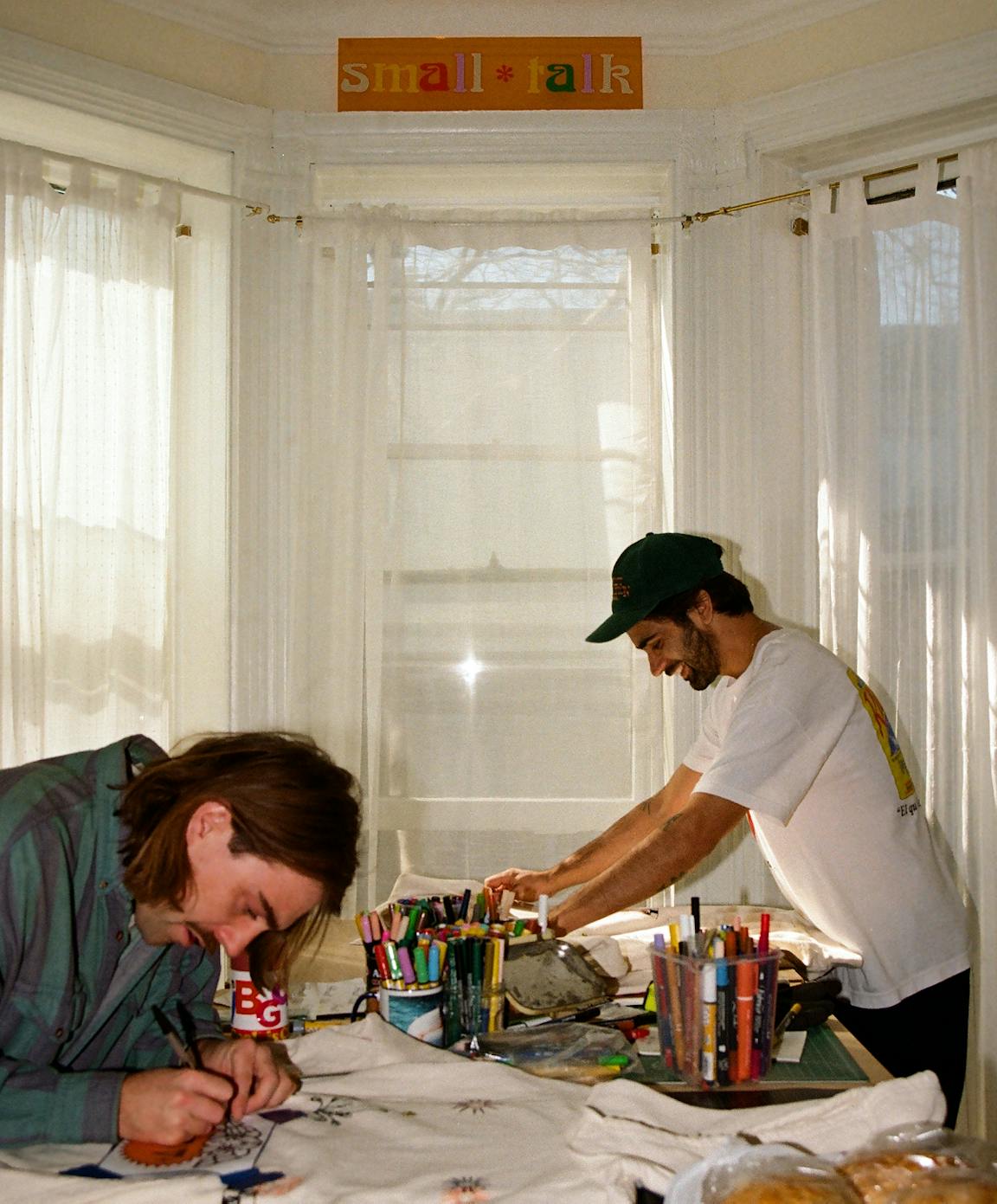Two masculine people work from a table full of art supplies.